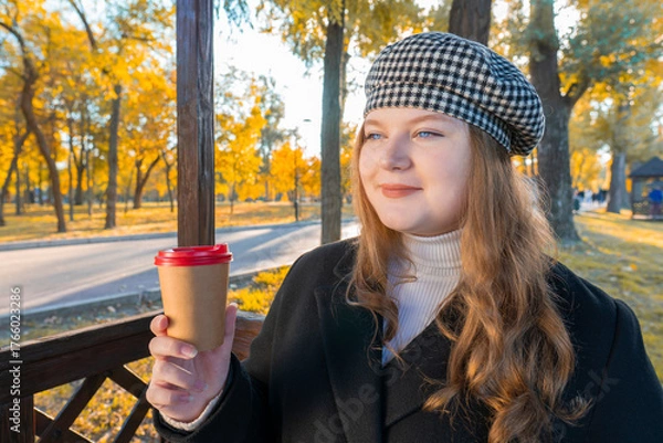 Fototapeta Close-up portrait of a smiling young woman in a checkered beret holding a paper coffee cup in an autumn park with golden foliage