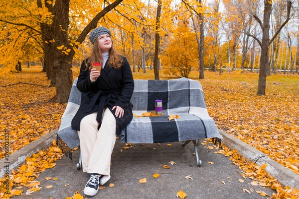 Fototapeta Young woman in a coat and beret sitting on a bench covered with a plaid blanket, enjoying a coffee in a beautiful autumn park
