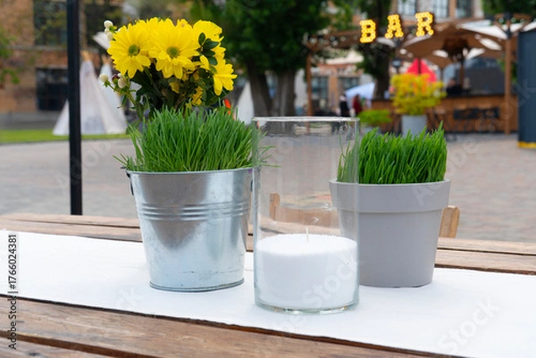 Fototapeta Cozy outdoor cafe table decoration featuring potted grass, yellow flowers, and a white candle in a glass holder with a bar sign blurred in the background