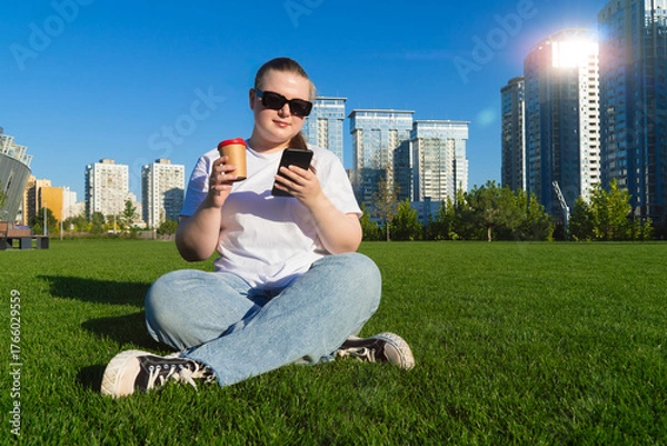 Fototapeta Young woman in sunglasses holding a phone and coffee cup while relaxing on green grass with modern skyscrapers in the background