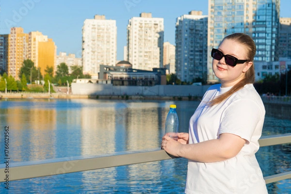 Fototapeta Portrait of a smiling young plus-size woman in sunglasses holding a plastic water bottle by a river with a modern cityscape behind her
