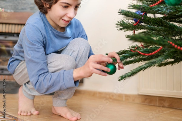 Fototapeta Child decorates the Christmas tree with red and shiny ornaments with excitement while wearing his pajamas