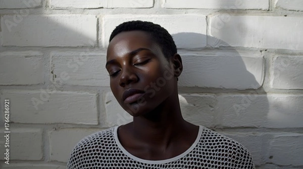 Fototapeta Artistic portrait of a serene young woman standing by a brick wall in soft sunlight, with gentle shadows highlighting her natural beauty, elegance, and introspective mood in minimal warm tones