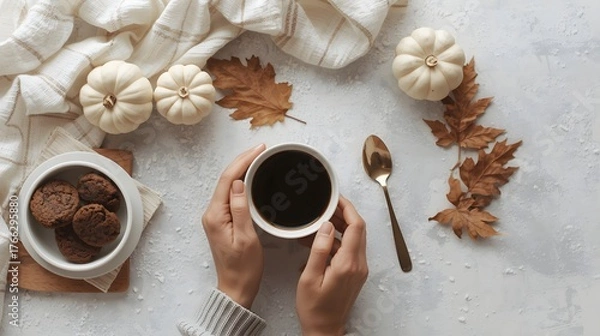 Fototapeta Cozy autumn flat lay with female hands holding a warm cup of coffee surrounded by mini white pumpkins, cookies, a golden spoon, and a dry maple leaf on soft fabric background in warm seasonal atmosphe