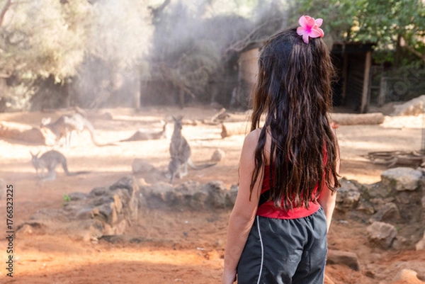 Fototapeta A young girl with long hair, wearing a red tank top and gray pants, stands quietly as she observes several kangaroos in a sunlit, dusty area of the zoo.