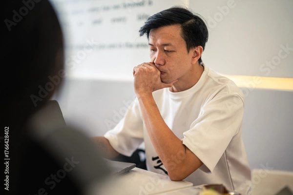 Fototapeta Asian man in a white shirt holding a notebook and pen while thinking deeply at a modern café table, with a laptop in front of him, showing a focused and contemplative expression in a bright setting