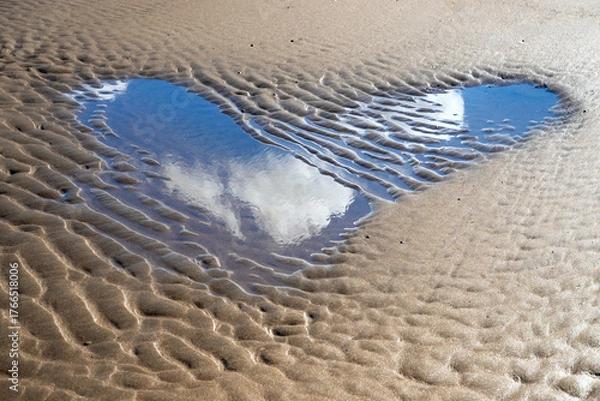 Fototapeta Heart-shaped small puddle on the North Sea beach of Norderney, reflecting white clouds in the blue sky. Tidal channel at low tide with typical sand structures that happen to form the symbol of love.