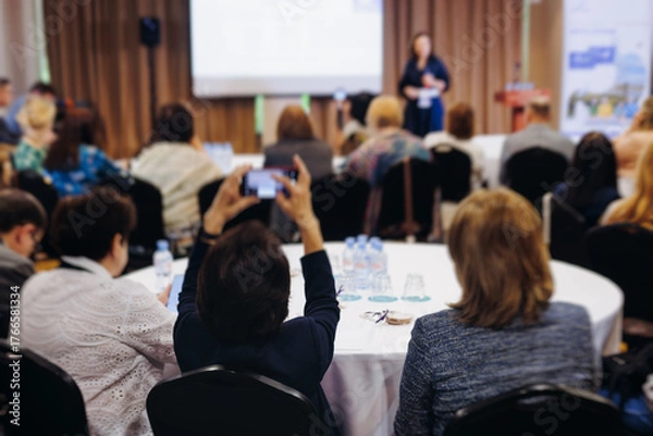 Fototapeta Female audience at the symposyum meeting, participants attendees in conference room hall listens to lecturer, group of women on a medical congress together listen to speaker on a stage at master-class