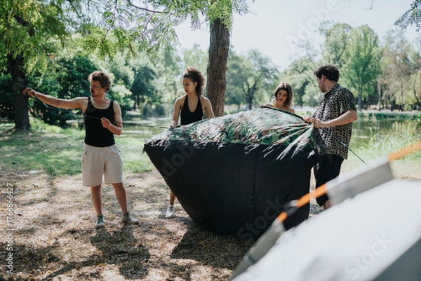 Fototapeta A group of friends works together to unfold and secure a large black tarp near a lake in a sunny park. They share teamwork and outdoor leisure during a casual camping trip.