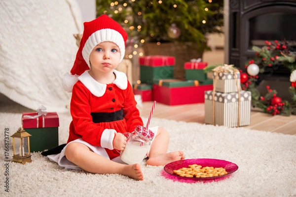 Obraz Kid in santa costume with milk and cookies on fluffy rug, surrounded by christmas tree, gifts, and fireplace.