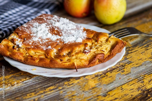 Fototapeta A homemade apple cake generously dusted with powdered sugar, presented on a rustic surface. The golden crust, soft texture, and visible apple pieces create a warm and inviting atmosphere, perfect for 