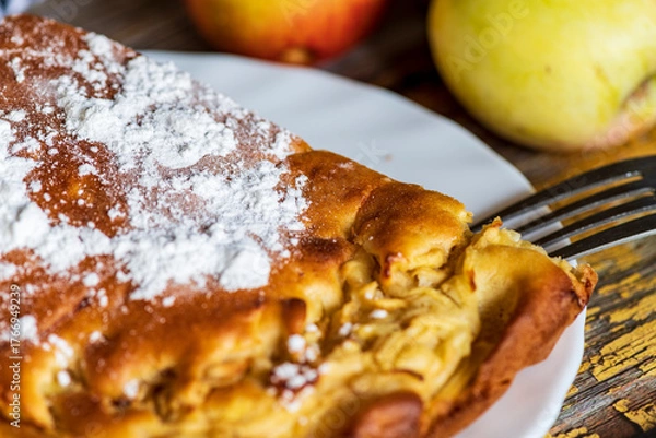Fototapeta A homemade apple cake generously dusted with powdered sugar, presented on a rustic surface. The golden crust, soft texture, and visible apple pieces create a warm and inviting atmosphere, perfect for 