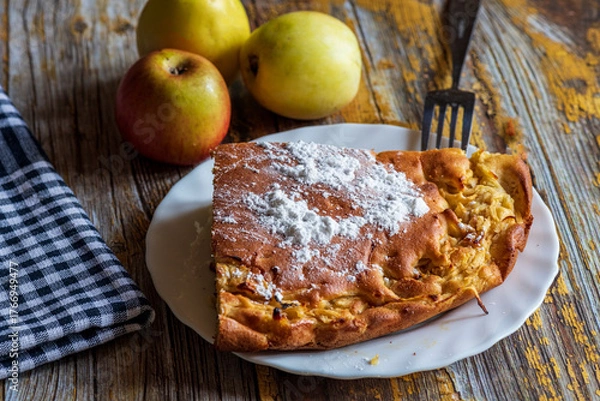 Fototapeta A homemade apple cake generously dusted with powdered sugar, presented on a rustic surface. The golden crust, soft texture, and visible apple pieces create a warm and inviting atmosphere, perfect for 