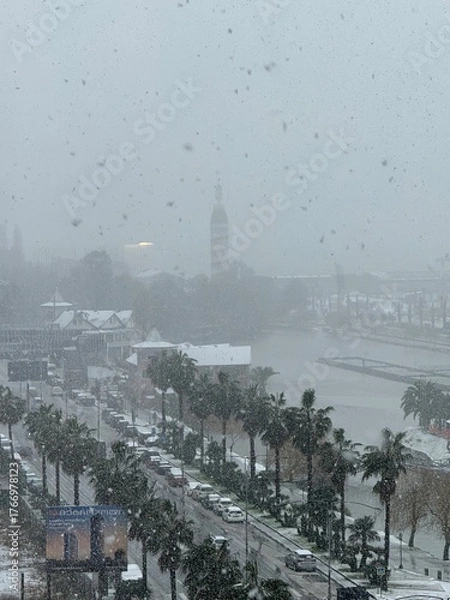 Fototapeta City street with cars and palm trees during heavy snowfall near a coastal tower. Used in travel, climate, and urban environment materials.