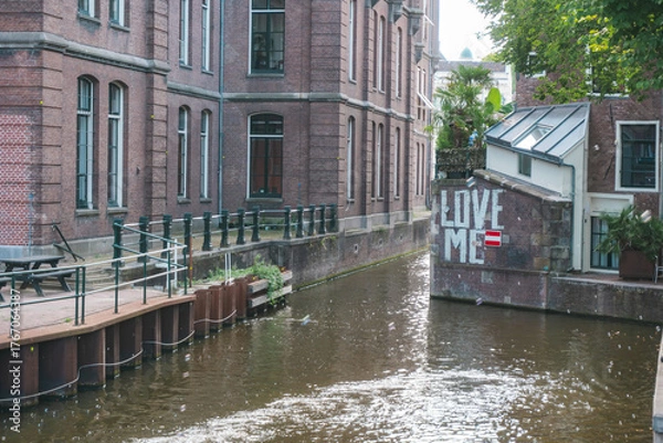 Fototapeta August 20, 2025, Amsterdam, Netherlands, view of a river on Grimburgwal Street with a sign reading “Love Me” in the background.