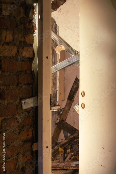 Obraz Partially open door reveals debris, collapsed beams, and crumbling walls inside an abandoned building illuminated by warm afternoon sunlight.