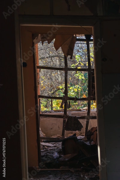 Fototapeta Interior of an abandoned house with a collapsed wall and shattered window, showing overgrown trees and sunlight outside through decay.