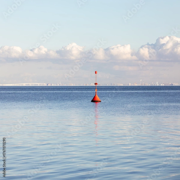 Fototapeta A bright red buoy floats in calm blue water under a soft cloudy sky, with a distant industrial coastline visible on the horizon.