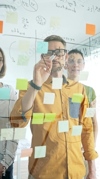 Fototapeta Business people team brainstorming, sharing strategy on glass whiteboard with colorful sticky notes, working collaboratively in bright office