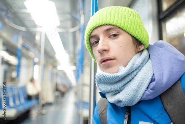 Fototapeta Portrait of a serious 12 year old boy in a green cap, white T-shirt and with a gray backpack in a subway car.	