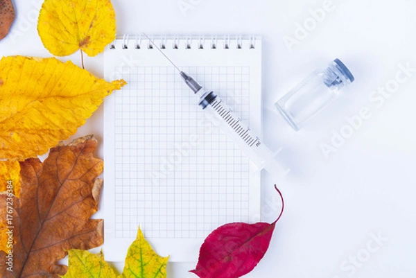 Fototapeta Vaccine and syringe are on table next to doctor's notebook and autumn leaves. Blank notebook for notes in doctor's office. Autumn vaccination..