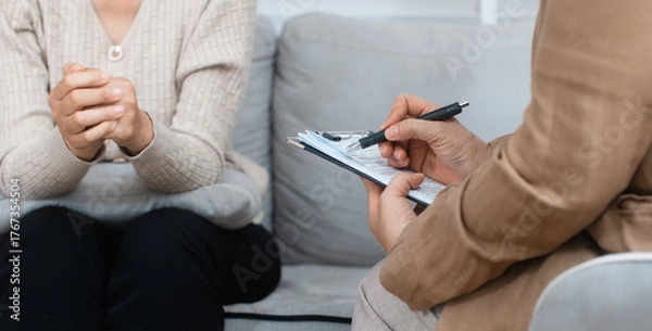 Fototapeta Close-up of psychologist hand writing, taking note about patient, listening to patient at psychotherapy session in office. Professional Psychologist consult with clipboard during treatment interview.