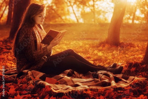 Obraz Beautiful young brunette sitting on a fallen autumn leaves in a park, reading a book