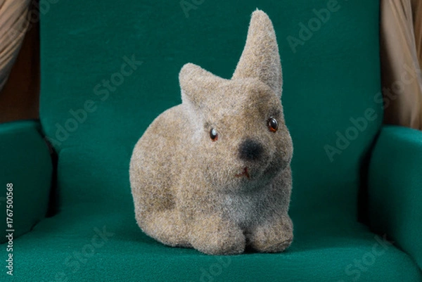 Fototapeta Adorable fuzzy brown bunny figurine with bright red eyes sits comfortably on a soft dark green upholstered chair, evoking warmth and charm.