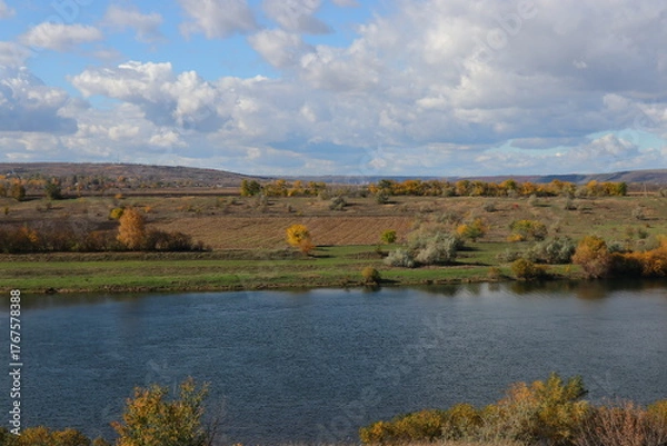 Fototapeta Peaceful autumn landscape with a calm river, colorful trees, and a grazing cow on a green hillside under a cloudy sky