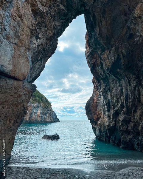 Fototapeta Scenic View Through Natural Rock Arch Overlooking Calm Ocean Waters and Rugged Coastline on a Cloudy Day. San Nicola Arcella, Calabria, Italy