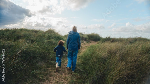 Fototapeta A mother and her child walk hand in hand through grassy sand dunes under a partly cloudy sky. The scene captures a peaceful moment of connection and exploration in nature, with soft natural light and 