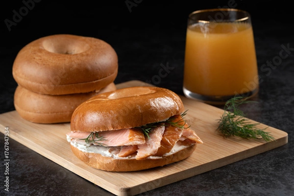 Fototapeta A sandwich with salmon and dill is on a wooden board next to a glass of juice. The sandwich is cut in half and placed on top of another half