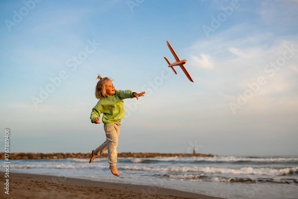 Obraz Smiling blond boy in green sweatshirt launching an orange toy airplane on the beach at sunset, looking at it as it flies above the sea.