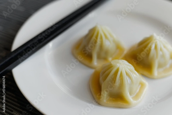 Fototapeta close-up of three delicate Xiao Long Bao (soup dumplings) with characteristic pleated tops, served on a white plate with black chopsticks