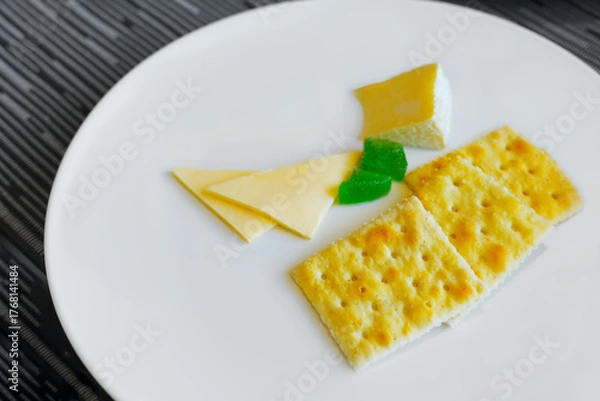Fototapeta A minimalist serving of square butter crackers and various slices of cheese, including a piece of brie or soft cheese, on a clean white plate