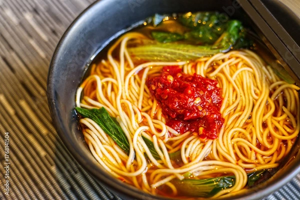 Fototapeta close-up of a dark bowl of hot noodle soup topped with a large dollop of vibrant red chili paste and green vegetables. 