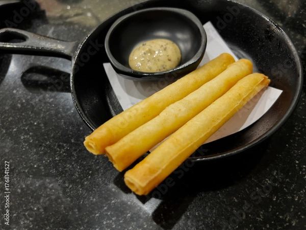 Fototapeta Three golden-brown fried spring rolls or cheese sticks served in a small black cast iron skillet with a side of creamy dipping sauce