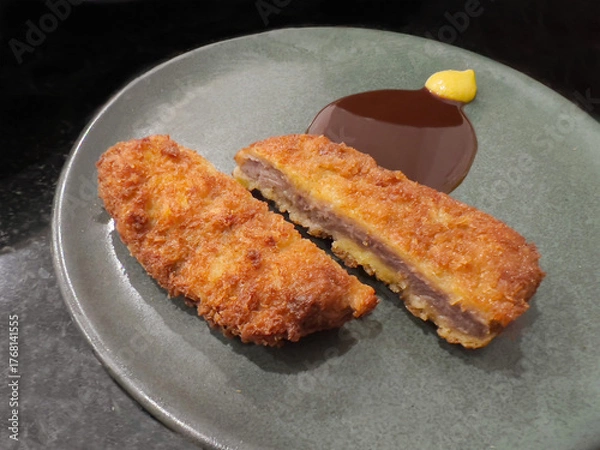 Fototapeta Two pieces of deep-fried, breaded meat cutlet (likely Beef or Pork Katsu), one sliced open, served on a modern grey-green plate. The dish is complemented by a thick brown sauce and a dollop of mustard