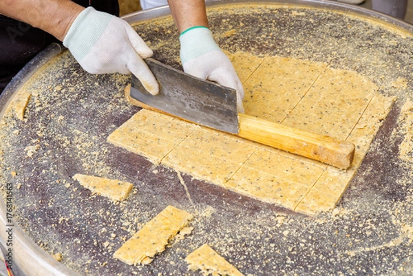 Fototapeta A close-up of a street vendor or baker cutting a large sheet of traditional yellow peanut brittle or sesame snack with a large knife on a round metal table.
