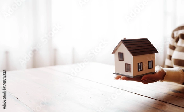 Obraz A person holding a small model house on their hand above a wooden table, symbolizing real estate, home ownership, and property investment.