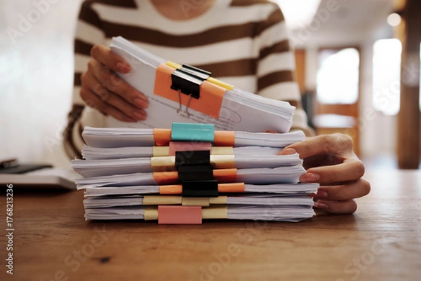 Obraz A person organizing stacks of documents with colorful binder clips on a wooden desk in a bright office environment.
