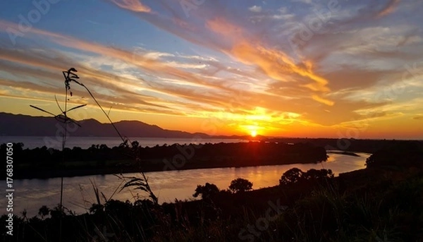 Fototapeta Sunset over a river, with vibrant orange and blue sky, grass silhouetted in the foreground, calm water reflecting light