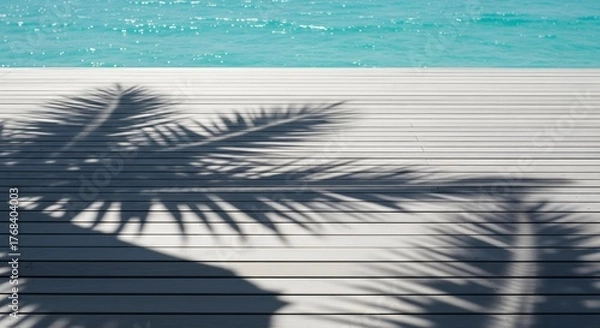 Fototapeta Palm tree shadow on a bleached wooden deck overlooking a vibrant turquoise ocean and calm sparkling water, creating a perfect tropical background