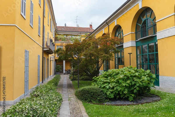 Fototapeta Greenery in the inner yard with pathwalks, historical stone bulding, yellow color, yearly XX century. Monza, Italy.