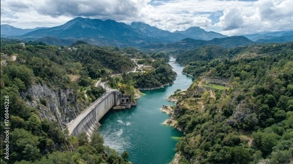 Fototapeta Scenic aerial view of a dam surrounded by lush greenery, clear river water, and majestic mountains under a cloudy sky in a serene landscape