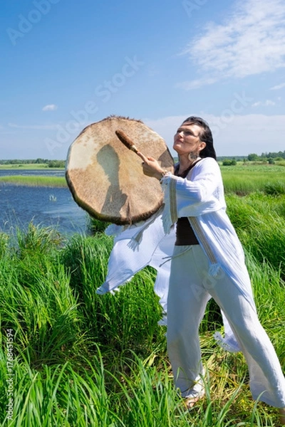 Fototapeta Middle-aged female shaman in the white dress drumming outdoors
