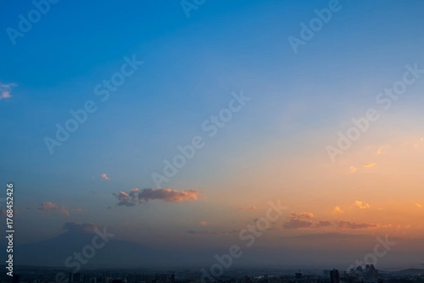 Fototapeta Distant view of Mount Ararat behind the city of Yerevan, Armenia at sunset. The sky fades from deep blue to warm orange, with soft clouds floating above the horizon