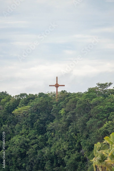 Fototapeta A large wooden cross standing atop a lush green hill surrounded by tropical trees under a cloudy sky