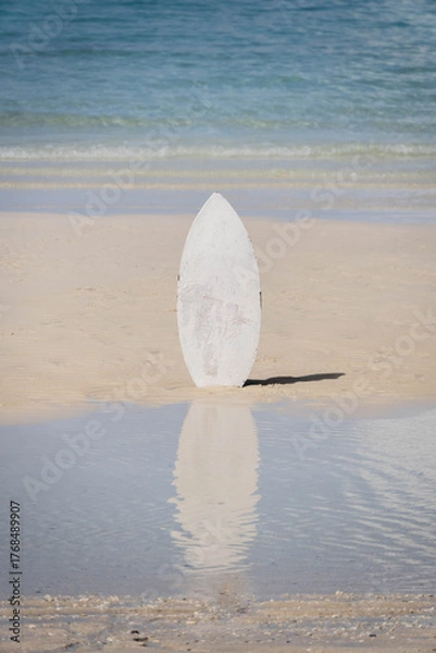 Fototapeta White surfboard standing upright on wet sandy beach with calm ocean waves in the background