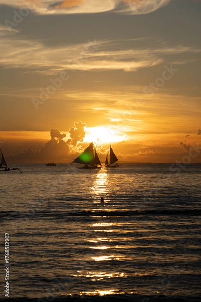 Obraz Sailboats gliding on calm sea during golden sunset with vibrant orange sky and reflections on water